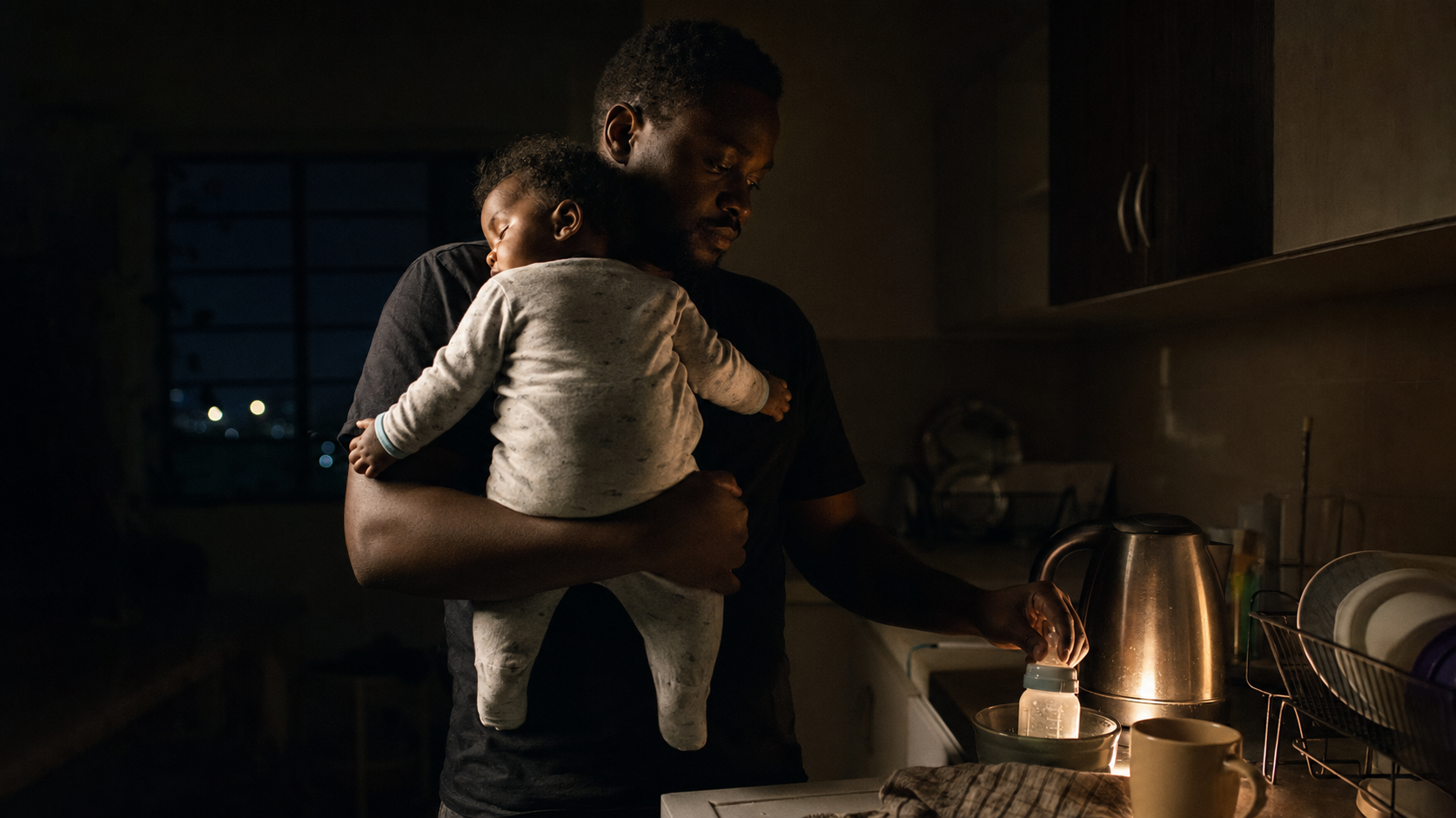 A father warming a bottle while holding his sleeping baby at night